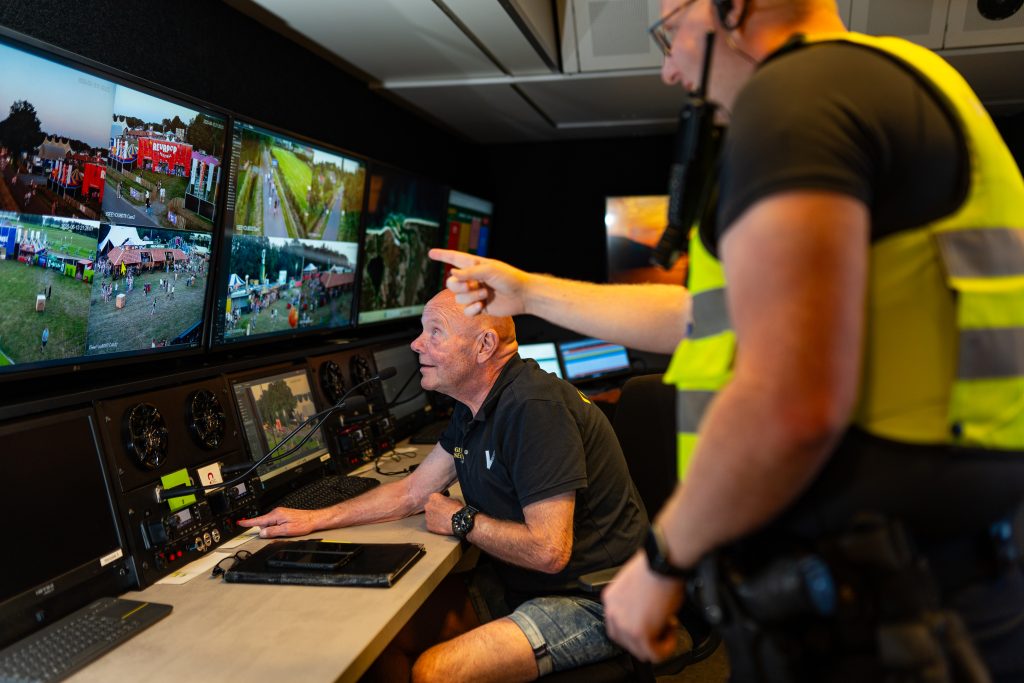 Coördinator en politie coördineren vanuit een mobiele controlekamer met meerdere camerabeelden van een festival op schermen.