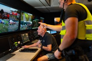 Coördinator en politie coördineren vanuit een mobiele controlekamer met meerdere camerabeelden van een festival op schermen.