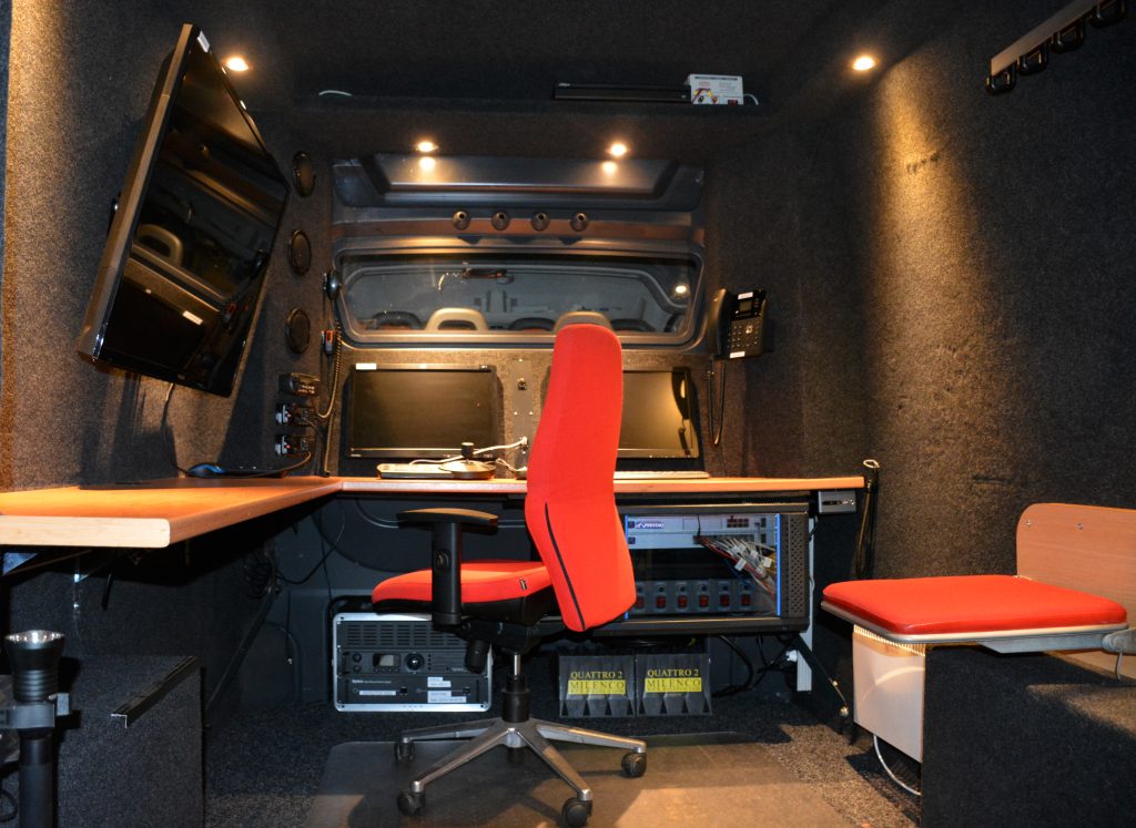 Binnenkant van een mobiele camera- en communicatie-unit met schermen en werkplek.
