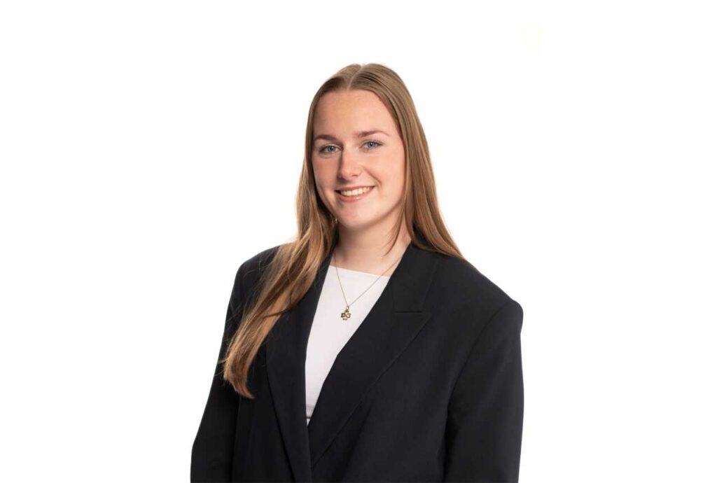 Zakelijke portretfoto van jonge vrouw in zwart colbert met witte top, glimlachend tegen neutrale achtergrond