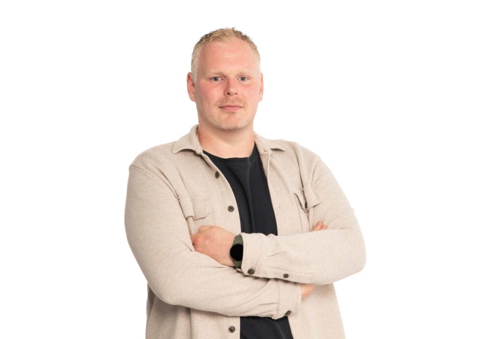 Professioneel portret van een man met gekruiste armen, gekleed in een licht overshirt en donker T-shirt tegen een neutrale achtergrond.