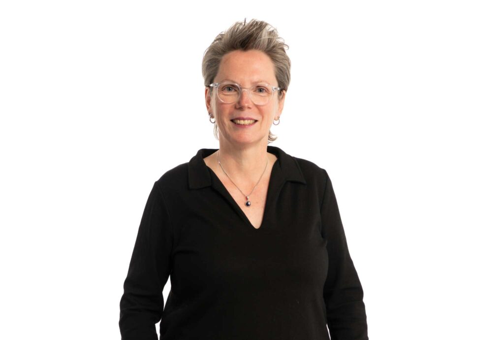 Professioneel portret van een glimlachende vrouw met bril en kort grijsblond haar, gekleed in een zwarte top tegen een lichte achtergrond.