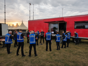 Beveiligingsteam krijgt een operationele briefing naast een mobiele commandowagen op een festivalterrein bij zonsopkomst.