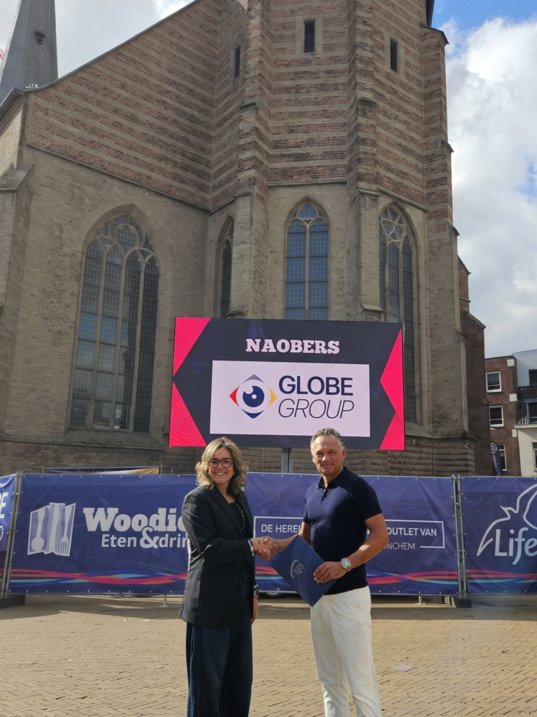 Twee vertegenwoordigers schudden elkaar de hand voor een kerk, met Globe Group zichtbaar op een groot scherm tijdens Naobers.