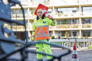 Verkeersregelaar in reflecterende veiligheidskleding met een verkeerskegel op zijn schouder, tijdens werkzaamheden in een woonwijk.